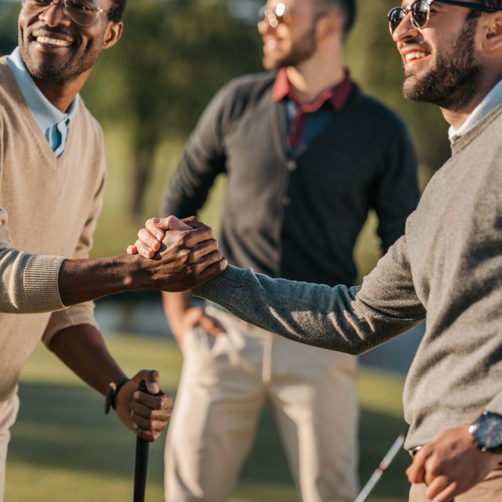 happy multicultural friends shaking hands while playing golf on golf course